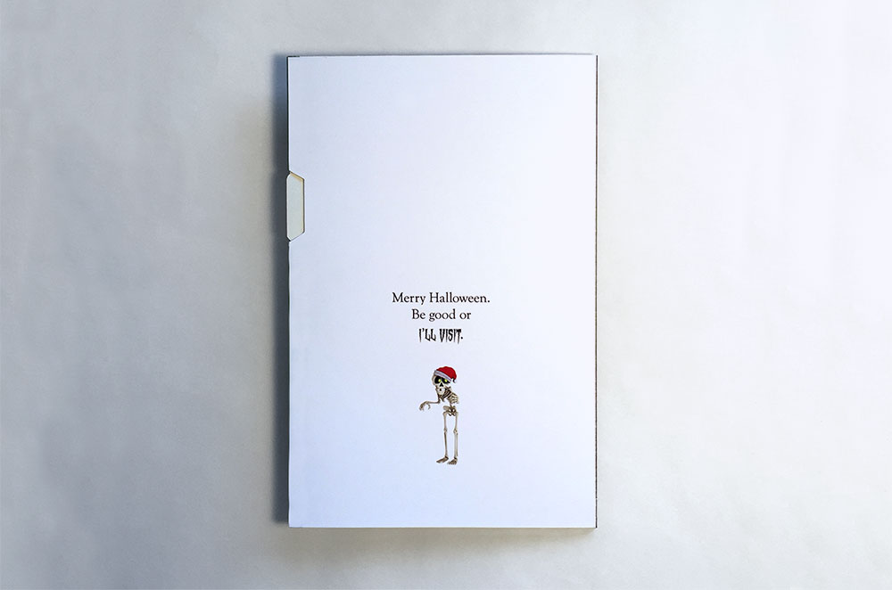 Back of card says, 'Merry Halloween. Be good or I'll visit.' above a skeleton wearing a Santa cap.